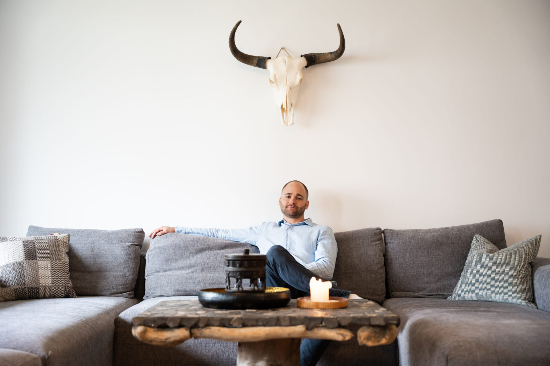 Konrad Mungenast sitzt auf einer grauen Sofagarnitur in einem modernen Wohnzimmer, an der Wand hängt ein Stierschädel, davor steht ein rustikaler Couchtisch aus Holz, perfekt in Szene gesetzt von Immobilienmakler Konrad Mungenast.